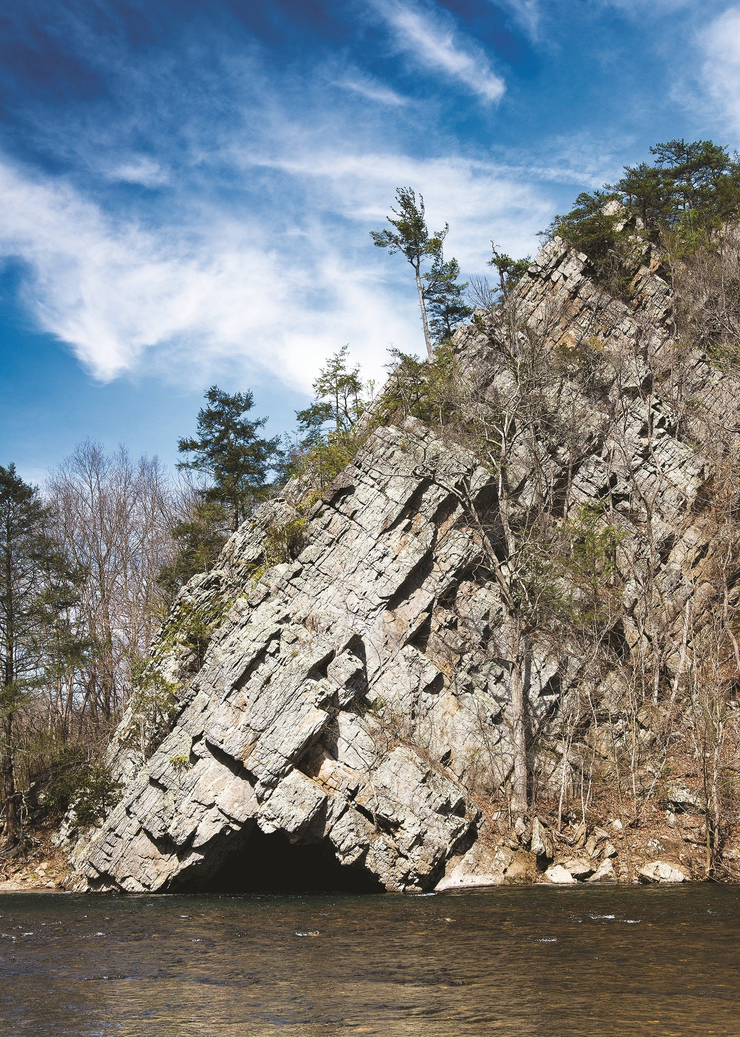 Rocky cliff with a cave entrance overhanging a body of water under a blue sky. West Virginia