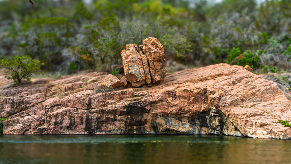 A smaller piece of red rock sits atop rocky ground of the same color. Water is in the foreground. Vegetation is in the background.