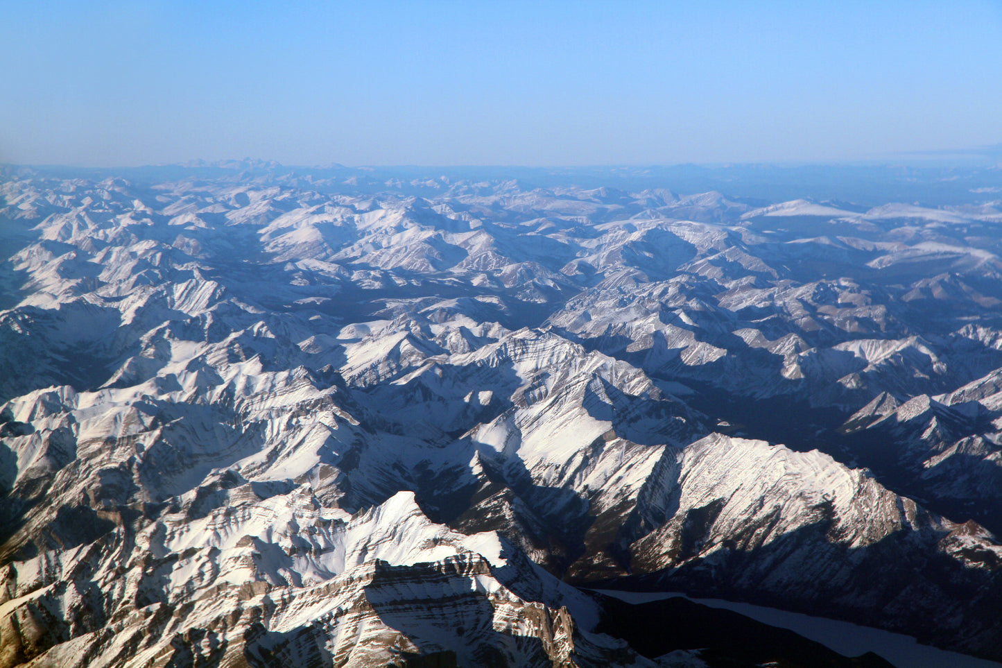 Snow-covered mountain tops are seen aerially. Foggy atmosphere and a blue sky is above. North America Cordilleran image.
