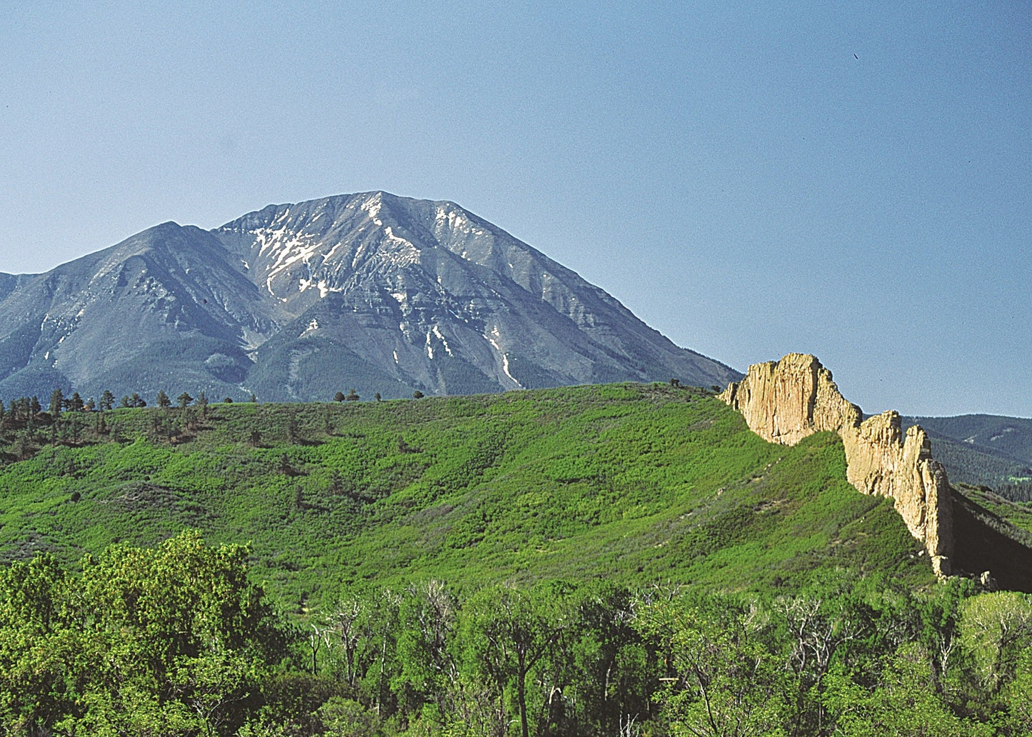 A scenic landscape photograph showing a large mountain with snow patches under a clear blue sky, with green hills and a rocky cliff in the foreground. Spanish Peaks, Colorado 