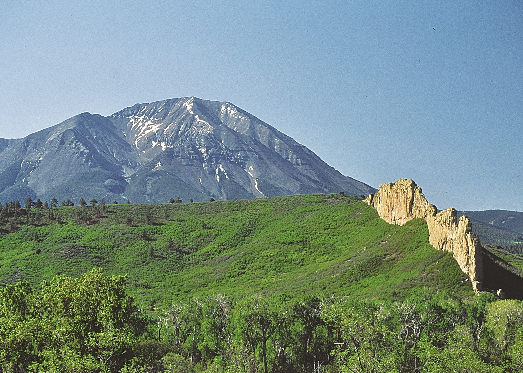 A scenic landscape photograph showing a large mountain with snow patches under a clear blue sky, with green hills and a rocky cliff in the foreground. Spanish Peaks, Colorado 