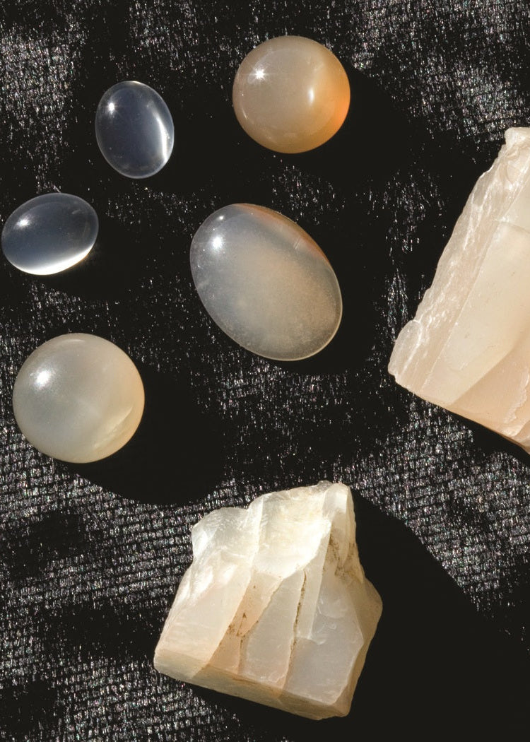 Five polished gemstone beads and two rough natural stone chunks arranged on a textured black fabric background.