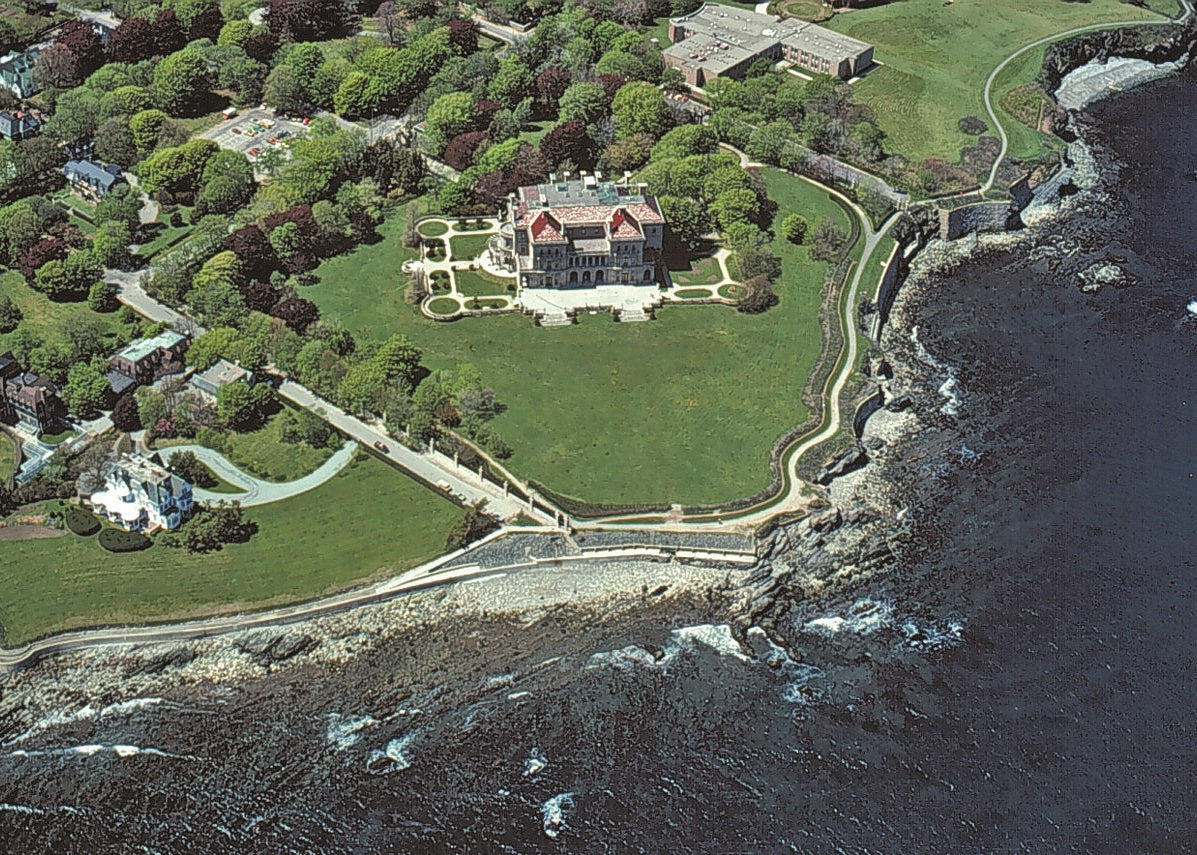 Aerial view of a large coastal estate with a green lawn, formal garden, mansion, rocky shoreline, and dark ocean water. Rhode Island