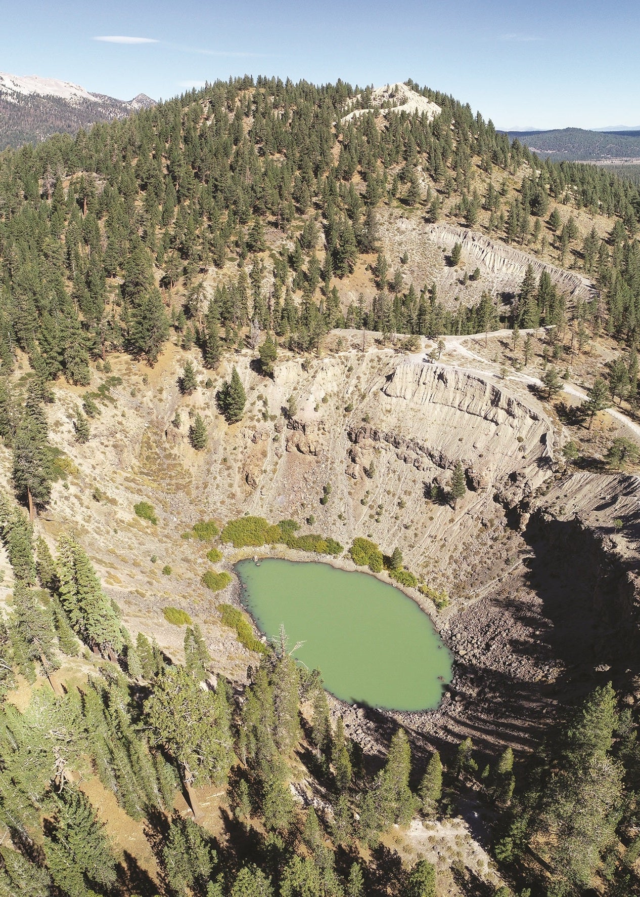 Aerial view of a rocky crater lake surrounded by dense evergreen forest and mountains under a clear blue sky. Death Valley, California 