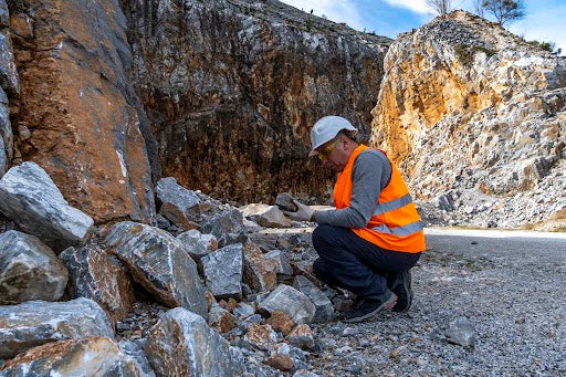 Person in a natural area surrounded by rock face wears orange safety vest, safety glasses, gloves, and a hard hat holds a rock larger than their hand and visually inspects it.
