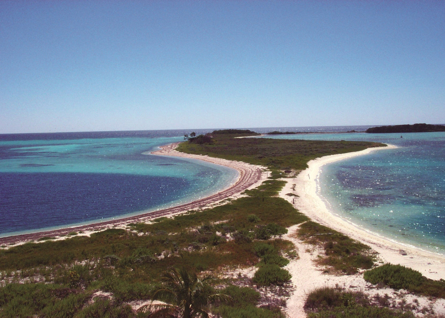 Aerial view of a tropical island with white sand beaches, green vegetation, and turquoise to deep blue ocean water under a clear blue sky. Florida