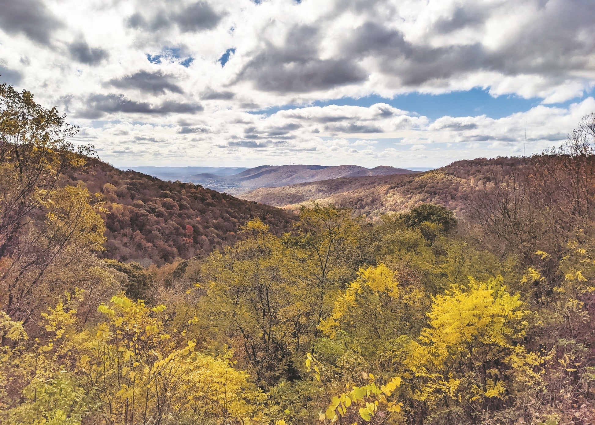 Mountain landscape with rolling hills covered in autumn trees, including bright yellow foliage, under a dramatic cloudy sky with patches of blue. View to the northeast toward the Jackson County Mountains from Monte Sano State Park, Alabama.