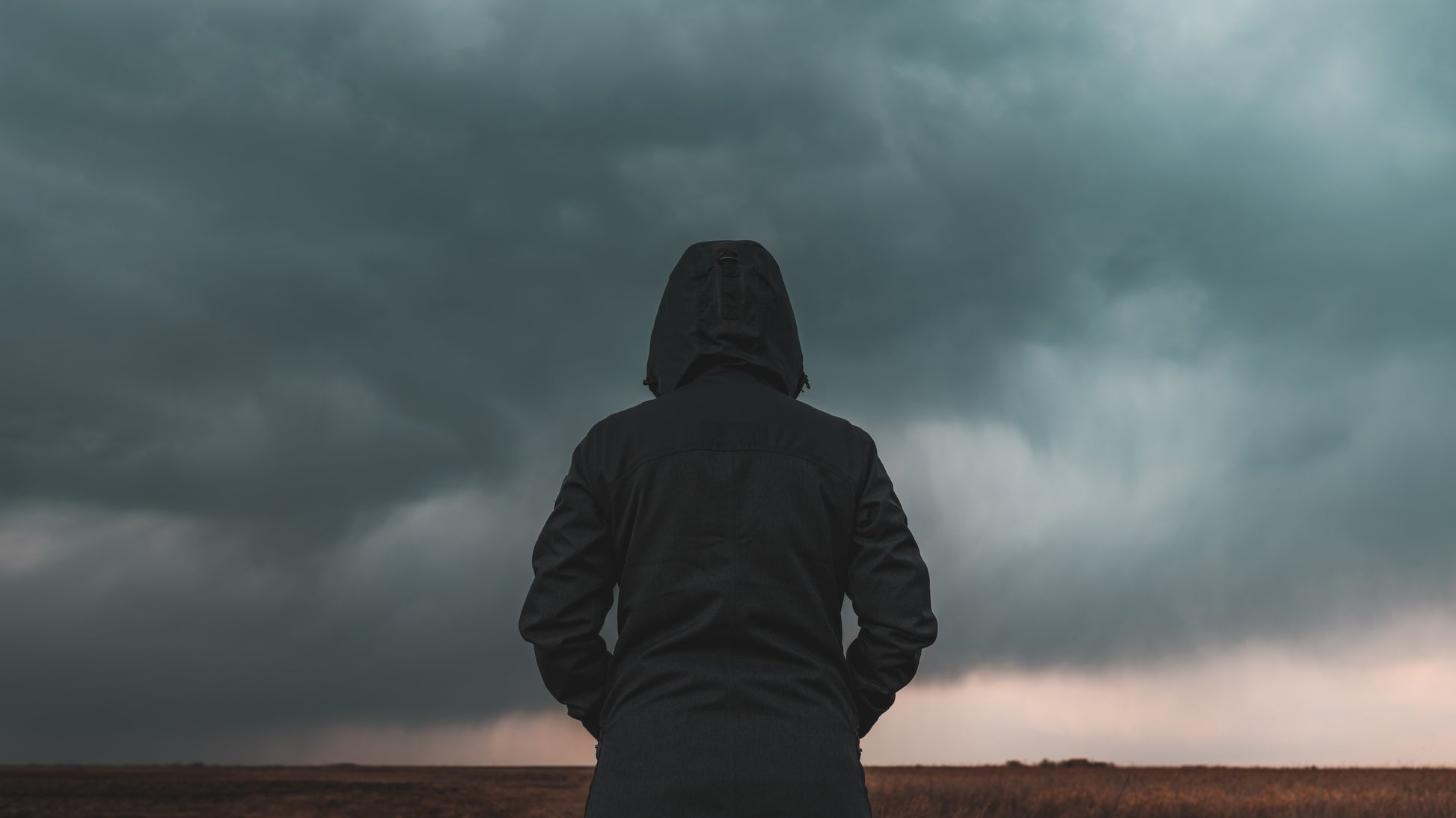 Person in a dark hooded jacket standing in front of a stormy sky. Climate Psychology image.