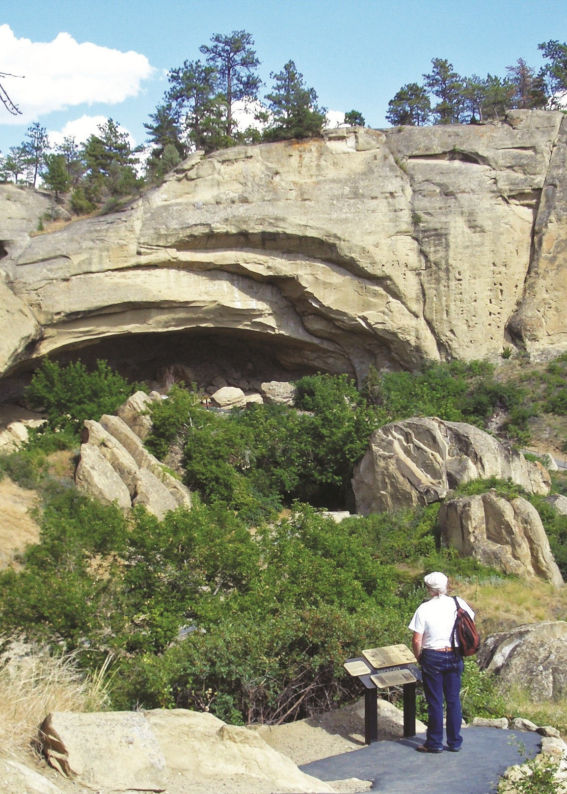 Person standing on a trail near large rock formations with trees in the background. Pictograph Cave, Montana