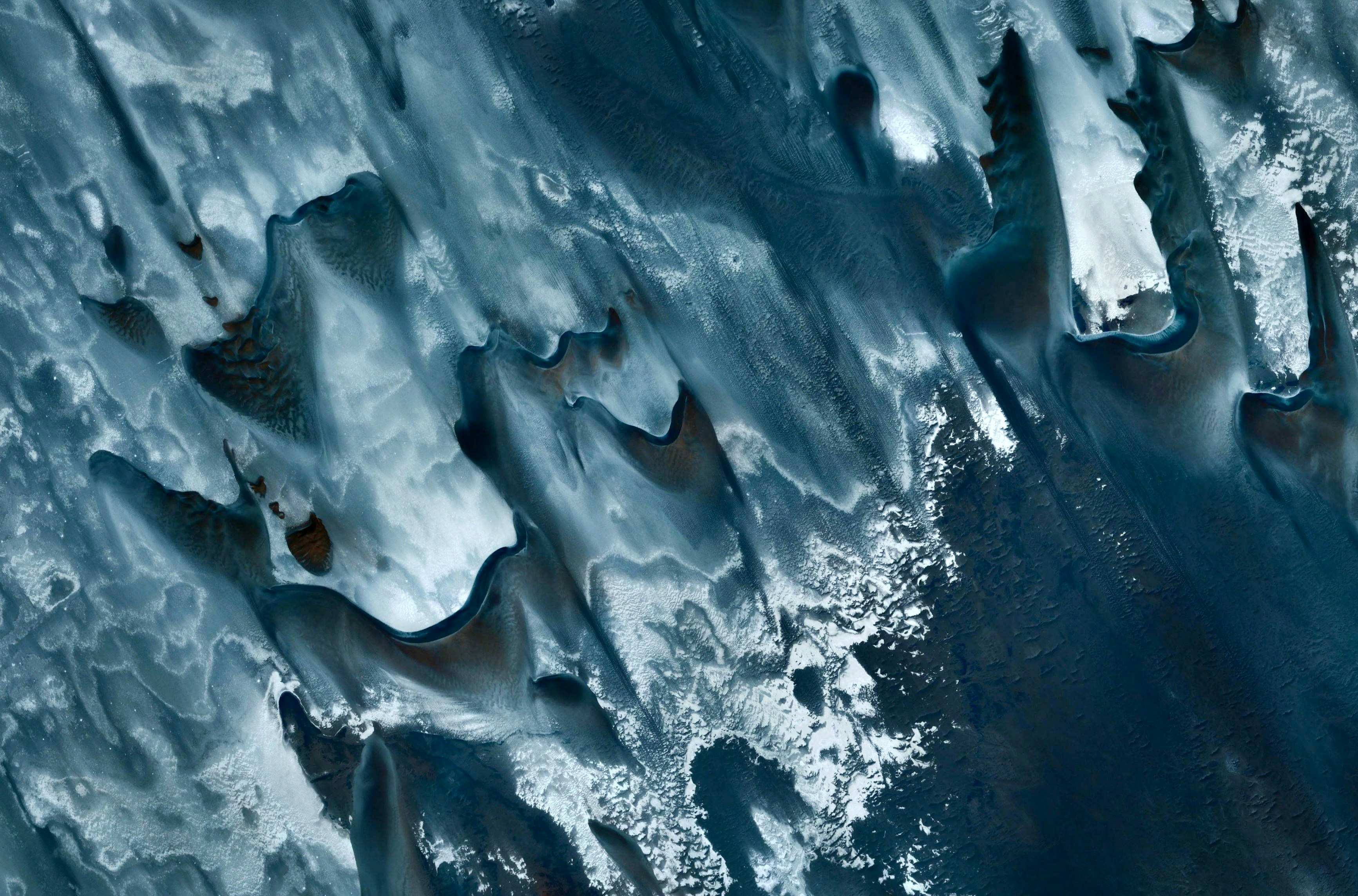 White and various tones of blue and gray make up this aerial photo of water, snow, and land (or sand) in this image captured from a plane.